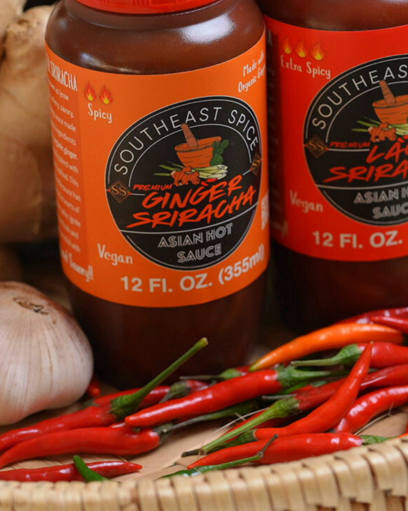 Bottles of Southeast Spice Ginger Sriracha and Lao Sriracha hot sauce surrounded by fresh garlic, ginger, and red chili peppers on a woven basket